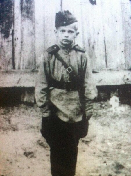 Сын полка Гена Абакумов стоит на фоне деревянного строения.